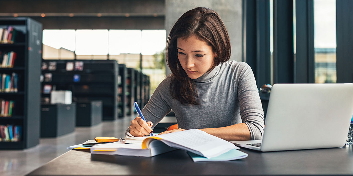 Student working in library writing in book with laptop on the table.