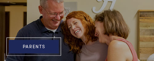 Parents hugging their daughter and laughing in dorm room.